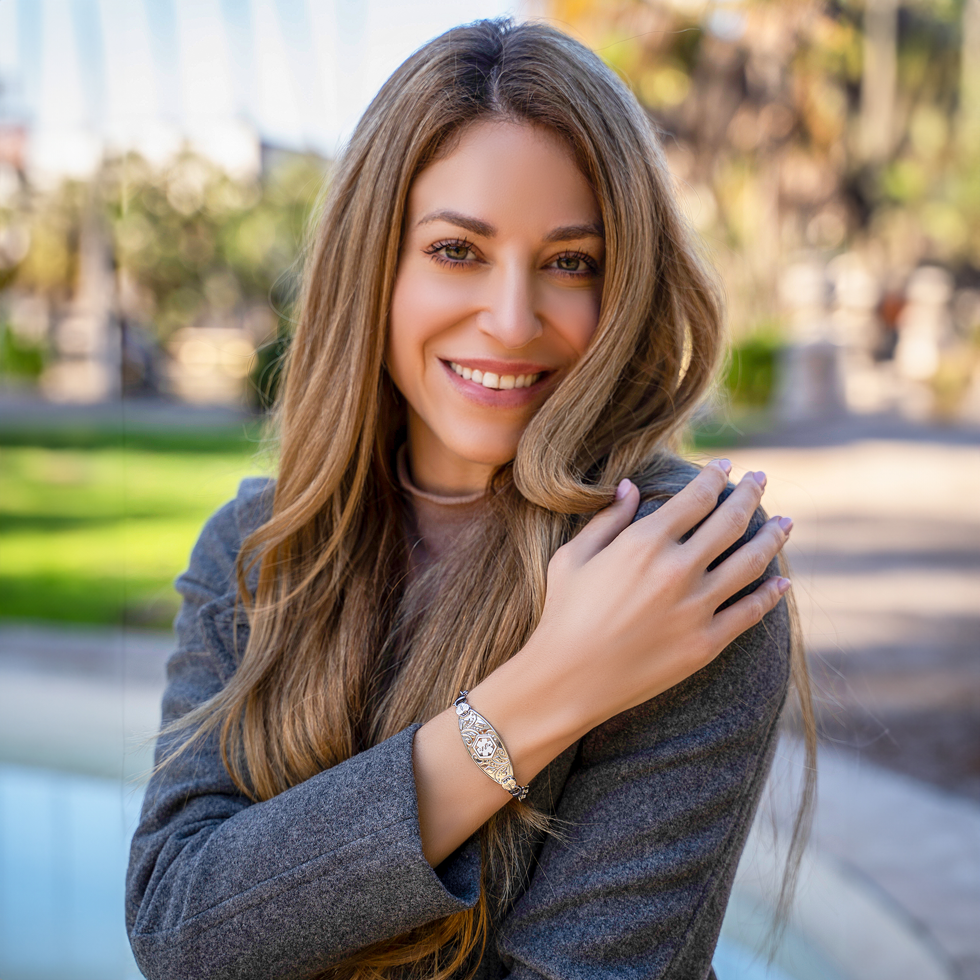 A portrait of a smiling woman with long, wavy light brown hair, wearing a grey blazer over a light-colored top. She cradles her right arm with her left hand, which rests on her shoulder, showcasing a silver bracelet with intricate metalwork and a central emblem. The background features blurred green and autumnal foliage, creating a warm and inviting outdoor setting.
