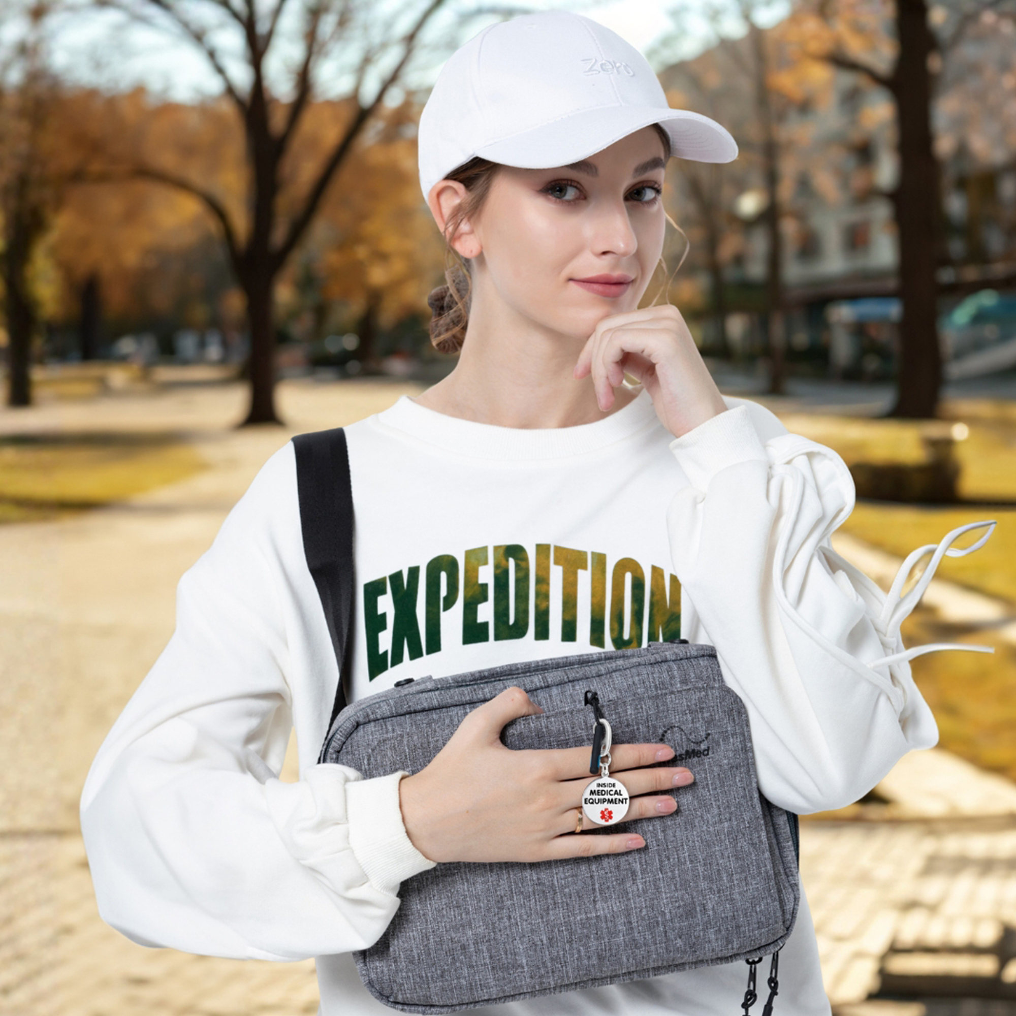 Young woman holding a grey ResMed bag with a circular medical alert keychain stating 'Inside Medical Equipment' and a red medical symbol, set against an autumn park background.