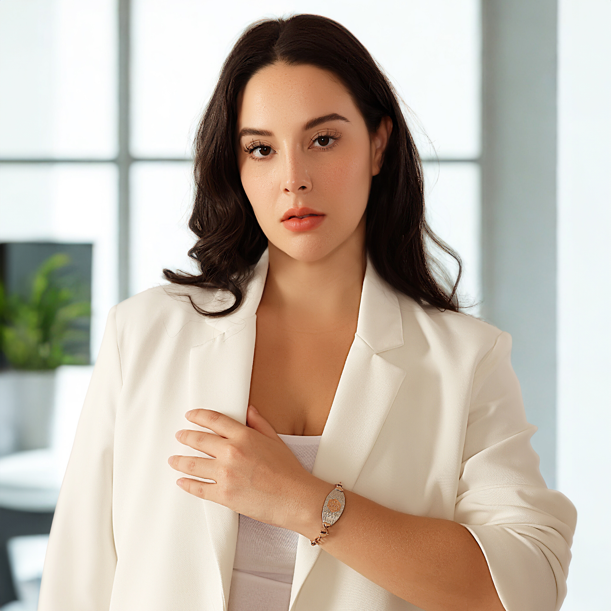 A woman with dark, wavy hair poses indoors against a bright, softly blurred background of large windows and a faintly visible green plant. She is wearing a white blazer over a white top and has a thoughtful expression while looking directly at the camera. Her left hand rests on her chest, showcasing a decorative bracelet on her wrist. The bracelet features an oval-shaped centerpiece with intricate rose gold and silver patterns, paired with a rose gold linked chain. The overall setting conveys a professional and elegant aesthetic.