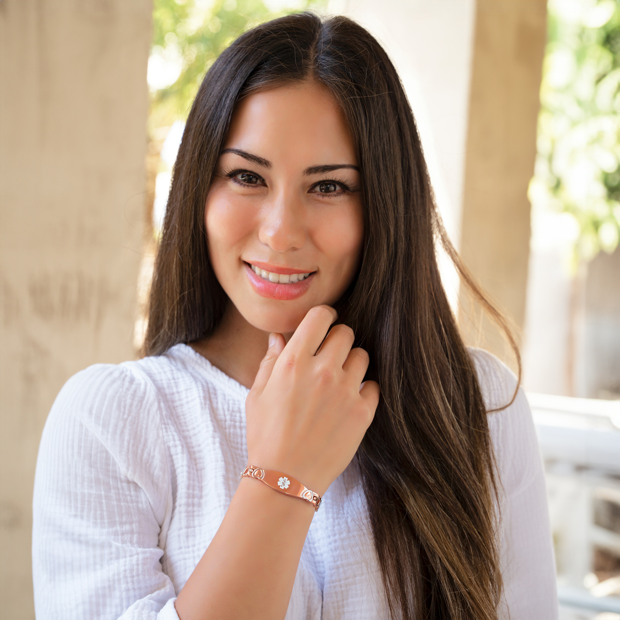 A smiling young woman with long dark brown hair and lighter highlights, wearing a white textured top and a rose gold medical alert bracelet. The bracelet features heart-shaped links and an oval plate with a white caduceus symbol and a star of life emblem. She is outdoors in a softly blurred background with greenery and sunlight, gently touching her chin with her left hand.