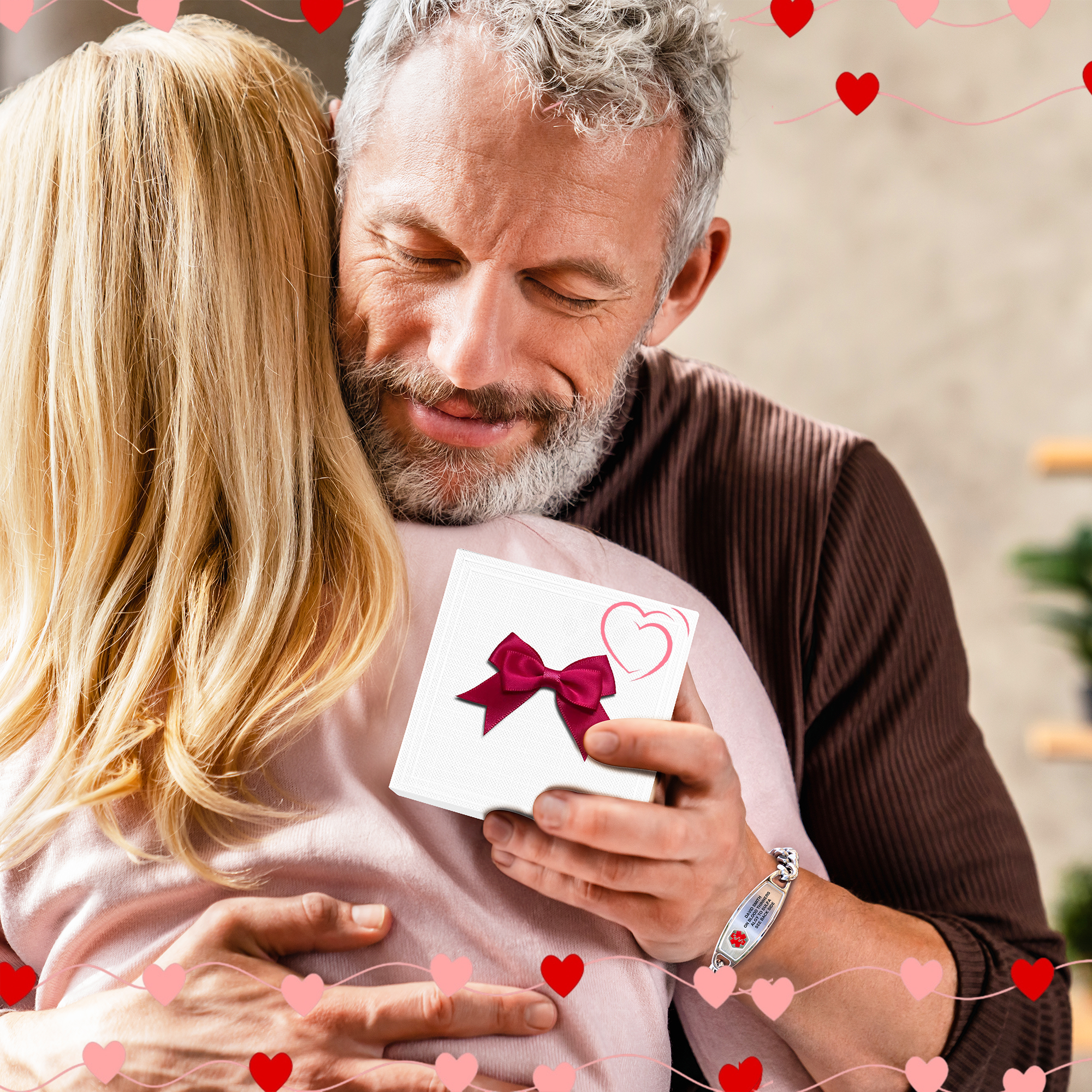 A close-up of a tender embrace between a man and a woman, surrounded by red and pink heart decorations. The man, with grey hair and a beard, wears a silver medical alert bracelet engraved with "DAVID SMITH," "ON BLOOD THINNERS," and "DIABETIC." He holds a small gift or card with a red ribbon and heart design against the woman's back. The woman, with long blonde hair, wears a light pink top and hugs him closely.