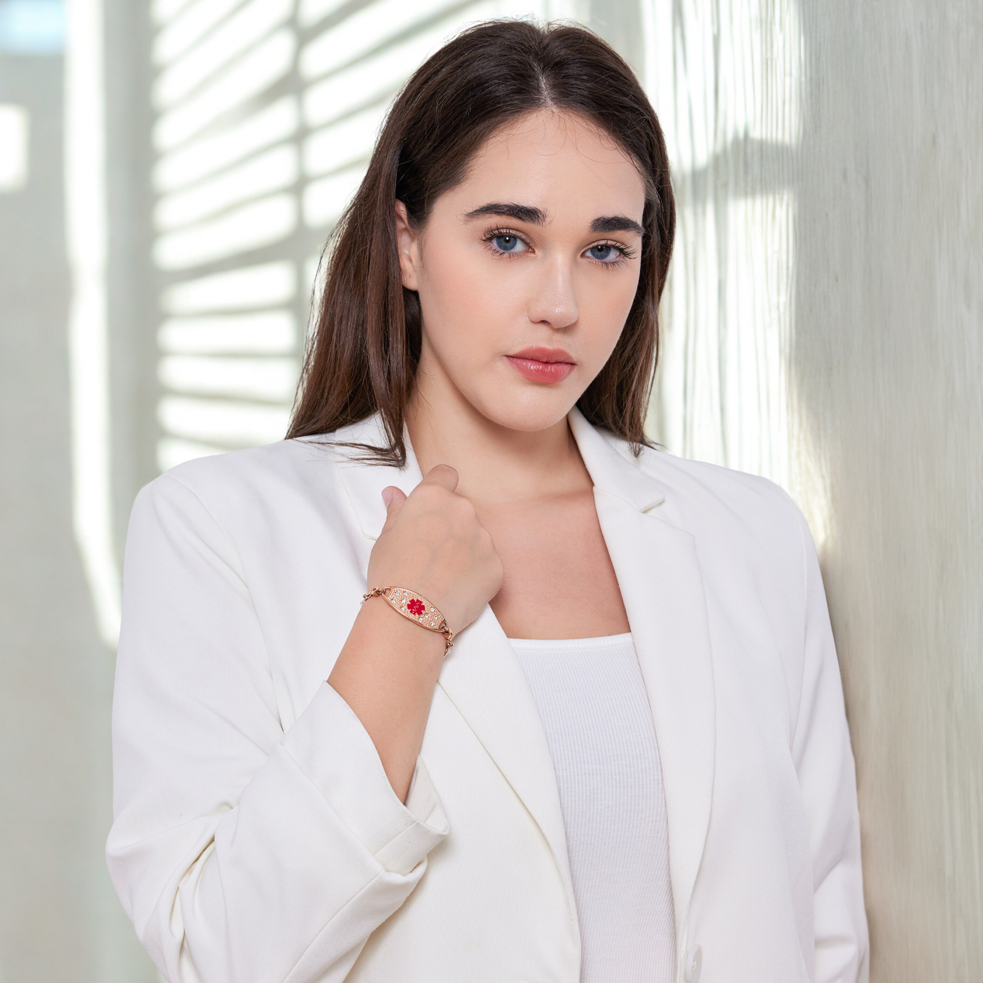 A young woman with long, dark brown hair and fair skin is wearing a white blazer over a ribbed white tank top. On her left wrist, she wears a rose gold medical alert bracelet with an oval charm, adorned with small clear stones and a red medical alert symbol in the center. She gently rests her left hand on her right upper arm, while standing against a softly lit indoor background with subtle shadows.