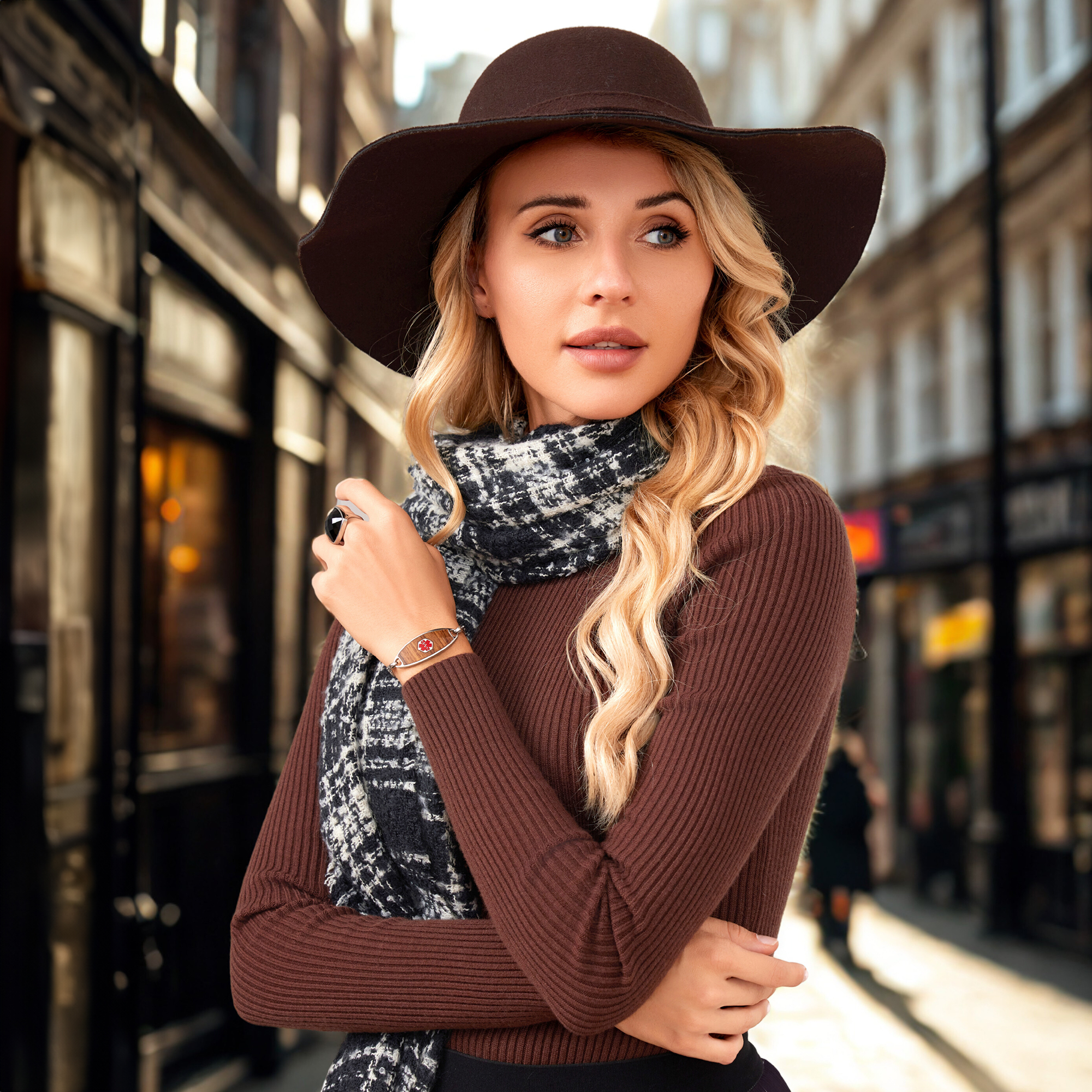 A fashionable young woman with long, wavy blonde hair stands in a blurred urban street setting. She wears a dark brown wide-brimmed hat, a black and white patterned scarf, and a ribbed brown long-sleeved top. On her left wrist, she has a metallic bracelet with a red medical alert symbol, and on her left index finger, she wears a ring with a large black stone. Her contemplative gaze is directed to the right, and the background features classic architecture and faint figures, emphasizing her as the focal point.