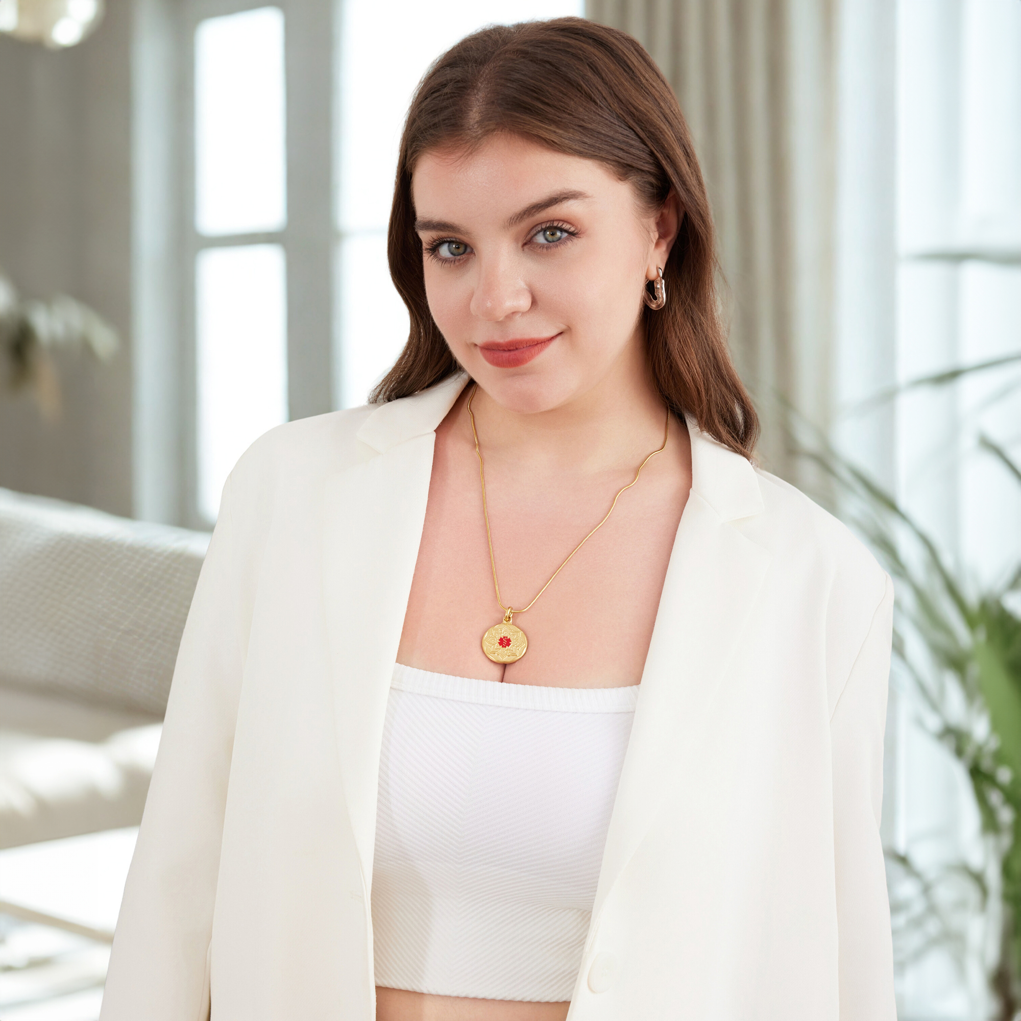 A young woman with long wavy brown hair and striking blue-green eyes poses indoors, wearing a white oversized blazer draped over her shoulders and a white ribbed crop top. She accessorizes with a gold hoop earring, a delicate gold chain necklace featuring a round pendant with a red floral emblem, and soft makeup. The background is softly blurred, showing hints of a modern living room with a sofa, a window with natural light, and green foliage.