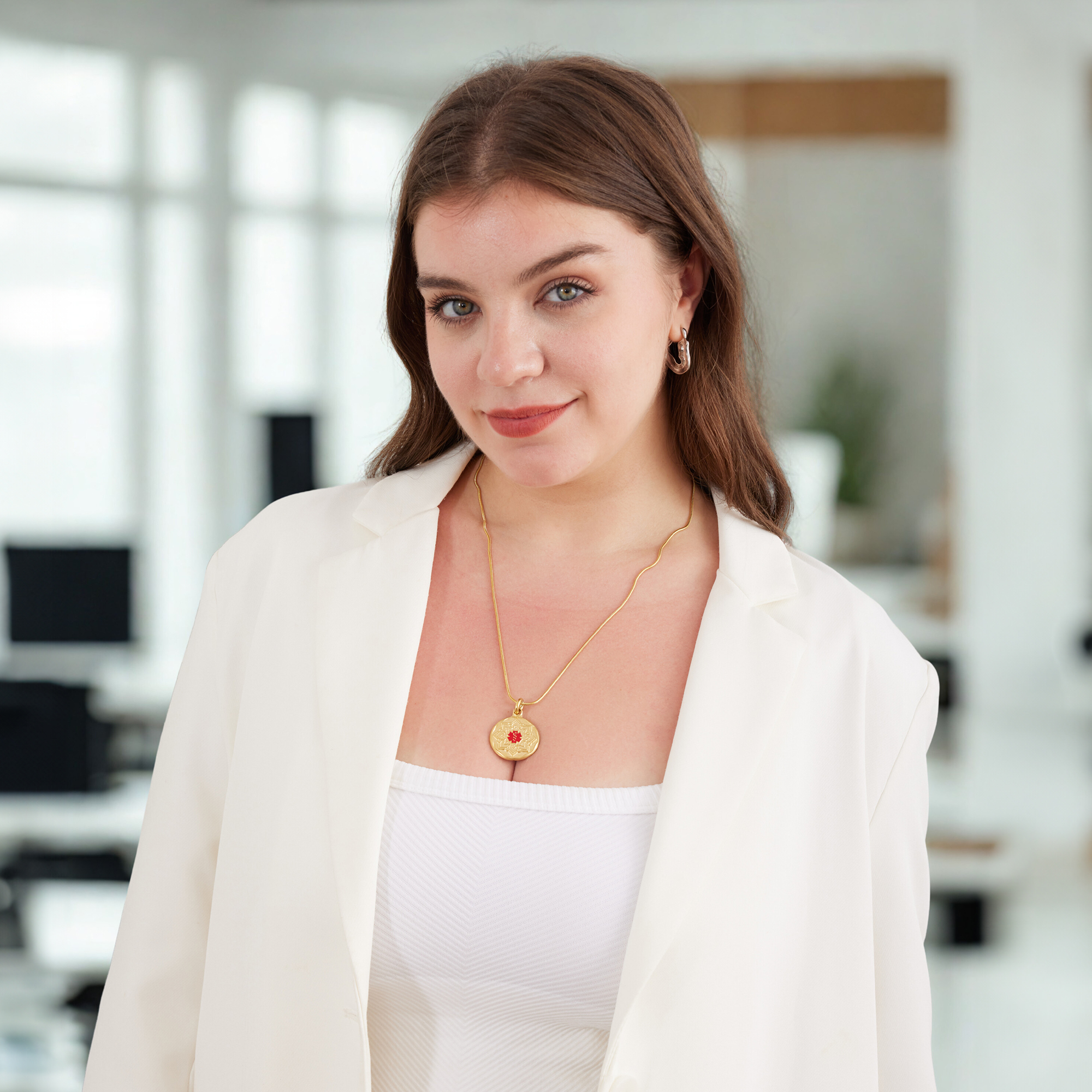 Portrait of a young woman with long brown hair, wearing a white blazer, a ribbed white top, a gold necklace with a red and gold pendant, and a gold hoop earring. She smiles softly while standing in a bright, blurred office setting.