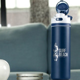 Navy blue water bottle with "SURF BEACH" logo, placed on a table next to decorative items, set against a soft, green couch background.