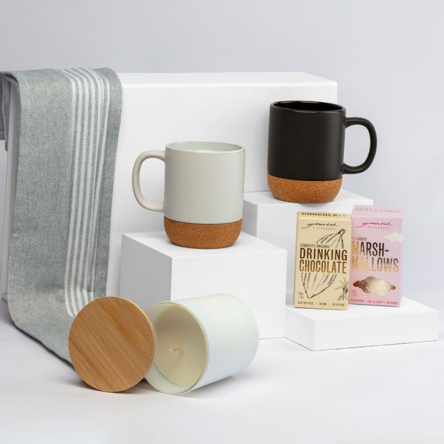 A collection of two mugs in grey and black with cork bases, a candle, and chocolate and marshmallow packets on white display.