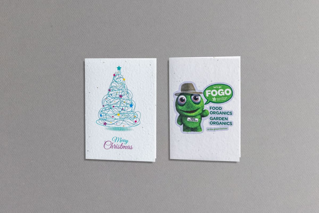 A large seed paper greeting card featuring a Christmas tree design and a green character illustration with a logo.