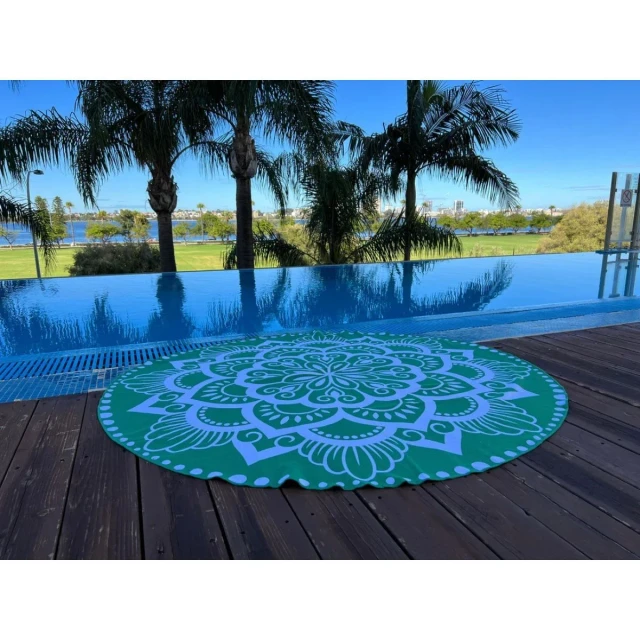 Round green sand-free rPET beach towel featuring a white mandala pattern, displayed on a wooden poolside deck with palm trees and a scenic waterfront view in the background.