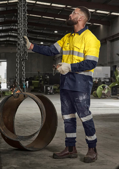 A man in a high visibility yellow and navy cargo pant with cuffs stands in an industrial setting, holding chains.