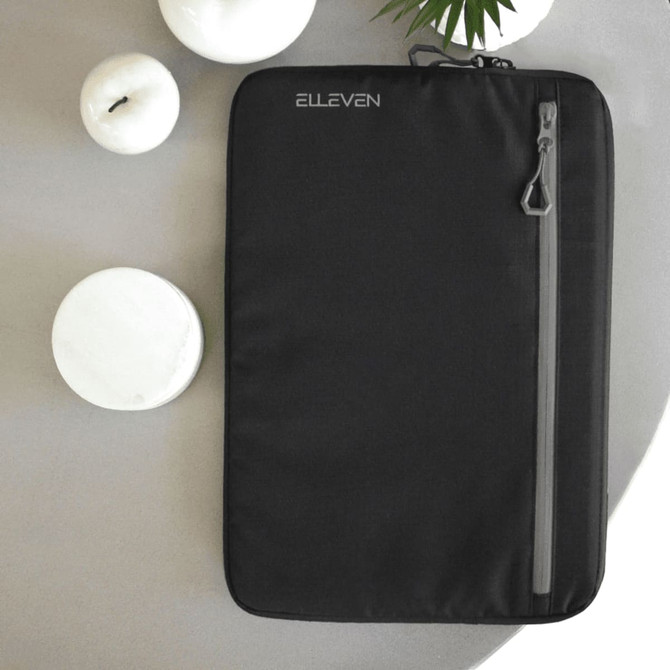 A black laptop sleeve featuring a front zipper pocket and a logo. Set against a neutral background with candles.