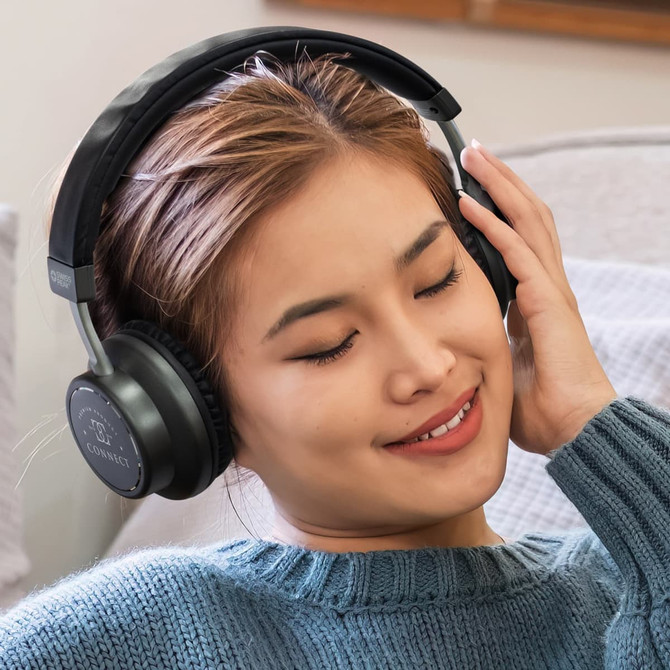 A pair of black wireless headphones worn by a woman with long hair, enjoying music in a cozy setting.