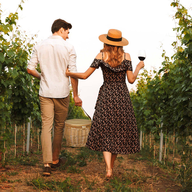A couple walks through a vineyard, the woman in a floral dress holding a wine glass and wicker basket. Sunlight filters through the vines.