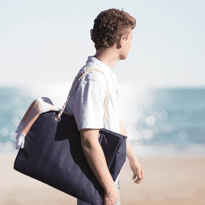 A waterproof navy beach bag with rope handles, carried by a person against a coastal background.