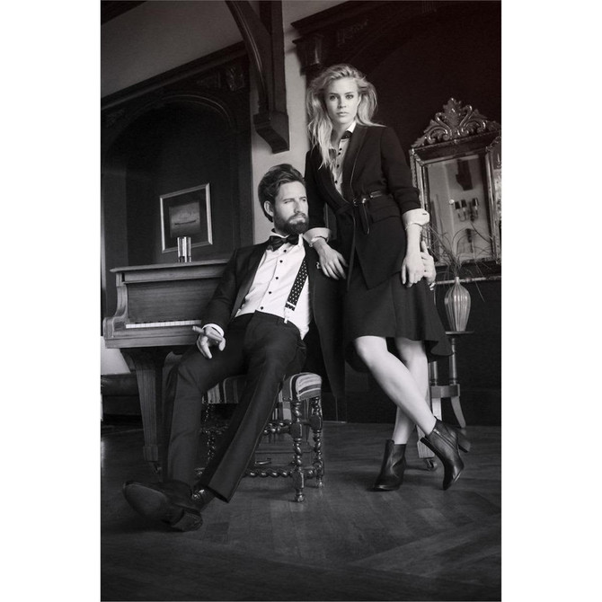 A woman in a black dress and high-heeled shoes stands next to a man in formal attire by a piano in a stylish room.