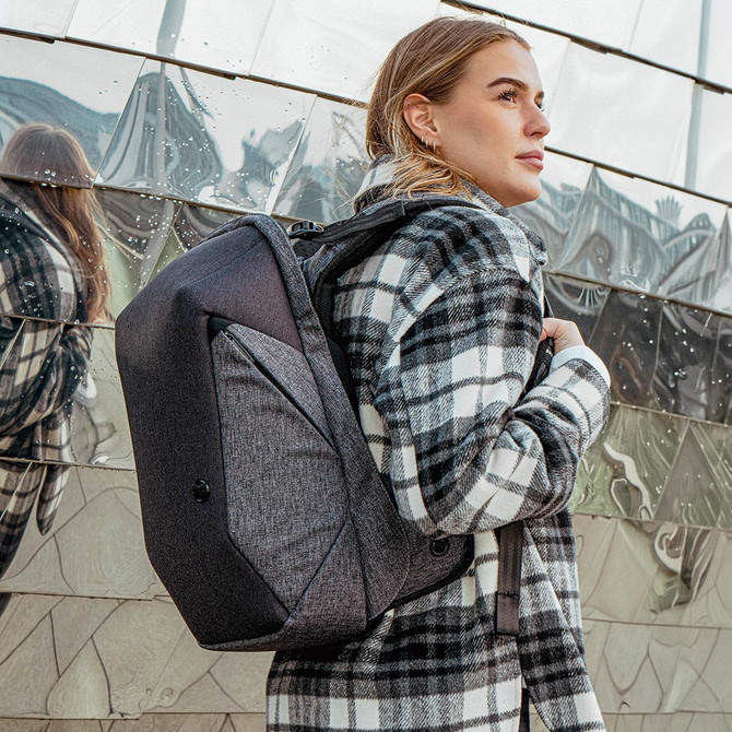 A grey backpack with a sleek design is worn by a woman in a checked coat, set against a reflective background.