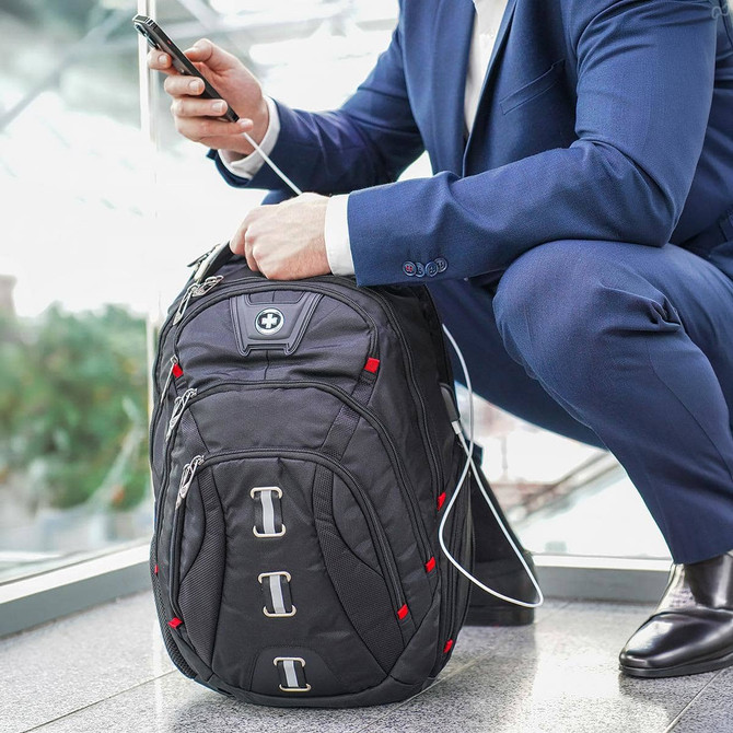 A black Swissdigital Pixel Backpack with red accents, positioned on the floor near a person using a phone.