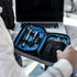 A black toiletry bag with a blue interior, containing pens and a small gadget, resting on a table beside a laptop.