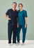 A pair of people wearing scrubs; one in navy jogger pants and the other in teal scrub top and pants. Both have a logo.