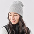 A grey knit beanie worn by a woman with long, wavy hair, paired with a dark vest. It features a logo.
