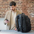 A black backpack sits on a table next to a man using his phone, with a charging cable connected to the bag.