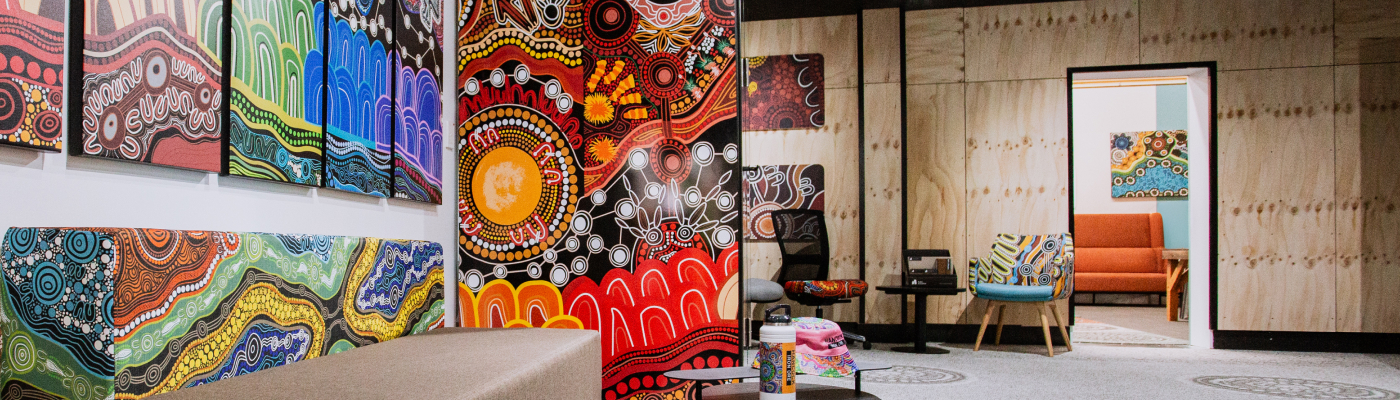 A room filled with vibrant abstract art on walls, featuring bold patterns and colors. A cozy seating area with colorful chairs and a sofa is visible in the background.