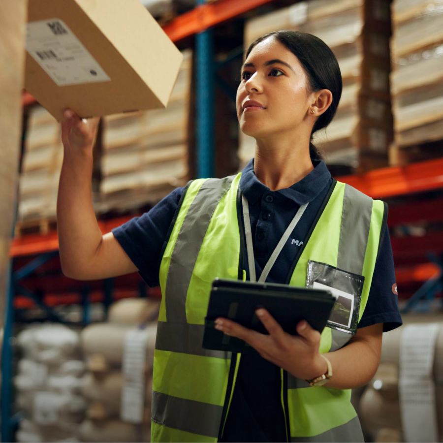 A woman in a warehouse wears a reflective vest and holds a tablet, reaching for a box