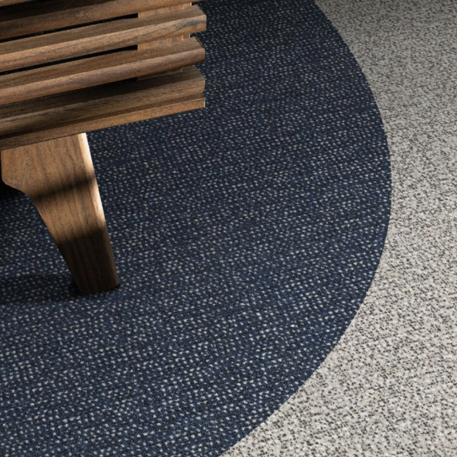 Corner of a wooden bench on a textured, circular blue carpet, set against a speckled gray floor. The contrast adds a modern and calming feel.