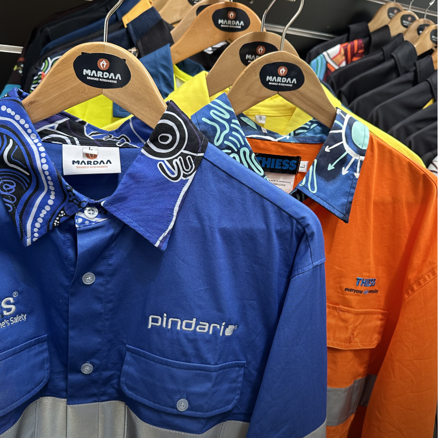 Colorful work shirts with artistic patterns hang on wooden hangers branded Mardaa. The shirts display vibrant blue and orange tones, conveying a lively atmosphere.