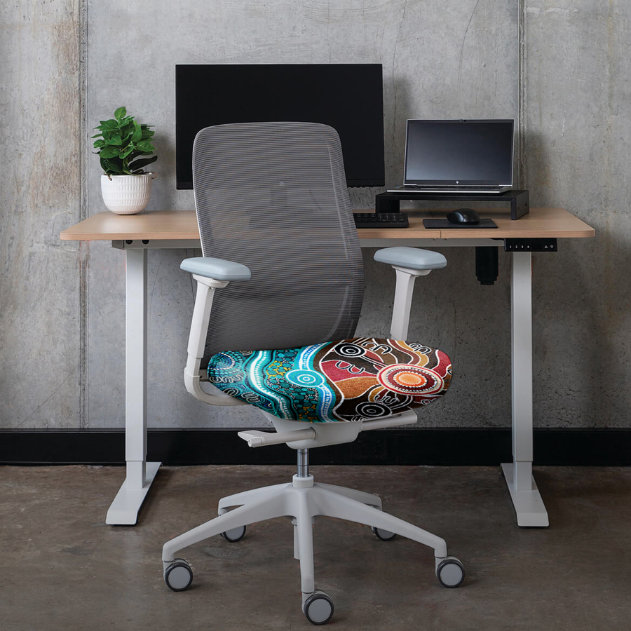 Modern office setup with a wooden desk, dual monitors, a potted plant, and a colorful ergonomic chair with a vibrant, abstract seat design.
