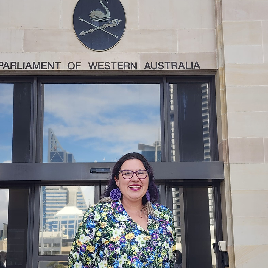Brooke-Louise O'Donnell stands outside the Parliament of Western Australia under a sign, with tall buildings reflecting behind her