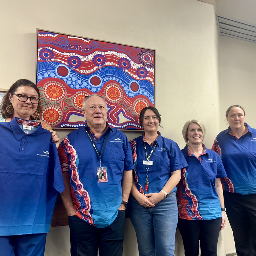 Five people in blue shirts with red and white Indigenous designs stand smiling in front of a vibrant, abstract artwork with similar patterns