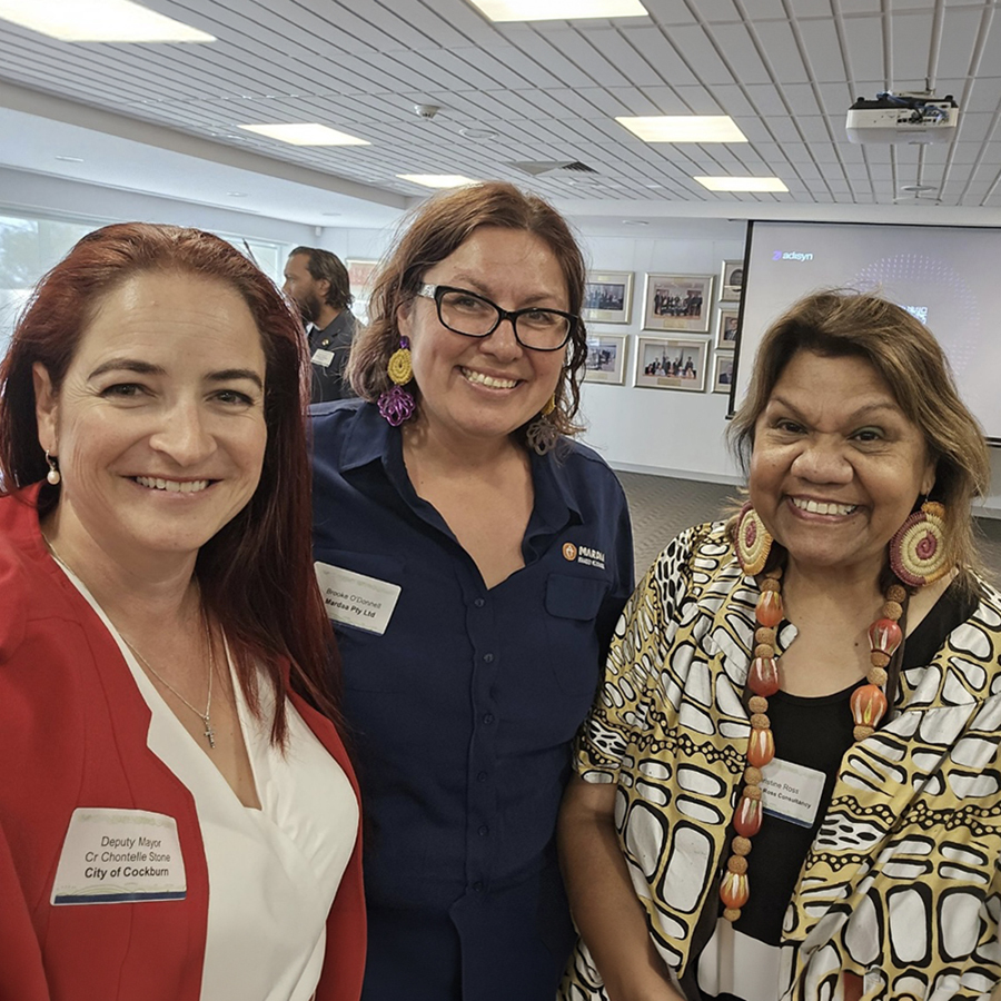Three women are smiling at an indoor event. One wears a red blazer, another a navy shirt, and the third a patterned blouse.