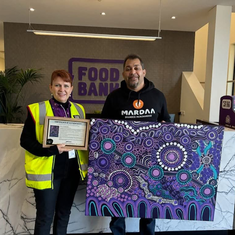Two people stand in a lobby with a "Food Bank" sign whilst one holds a certificate, the other an abstract art piece in purple and blue tones