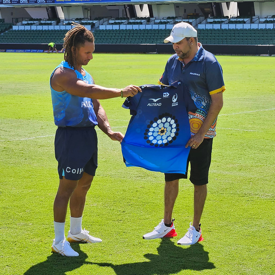 A man in sports attire presents a blue jersey with circular patterns to another man in a cap on a sunny sports field