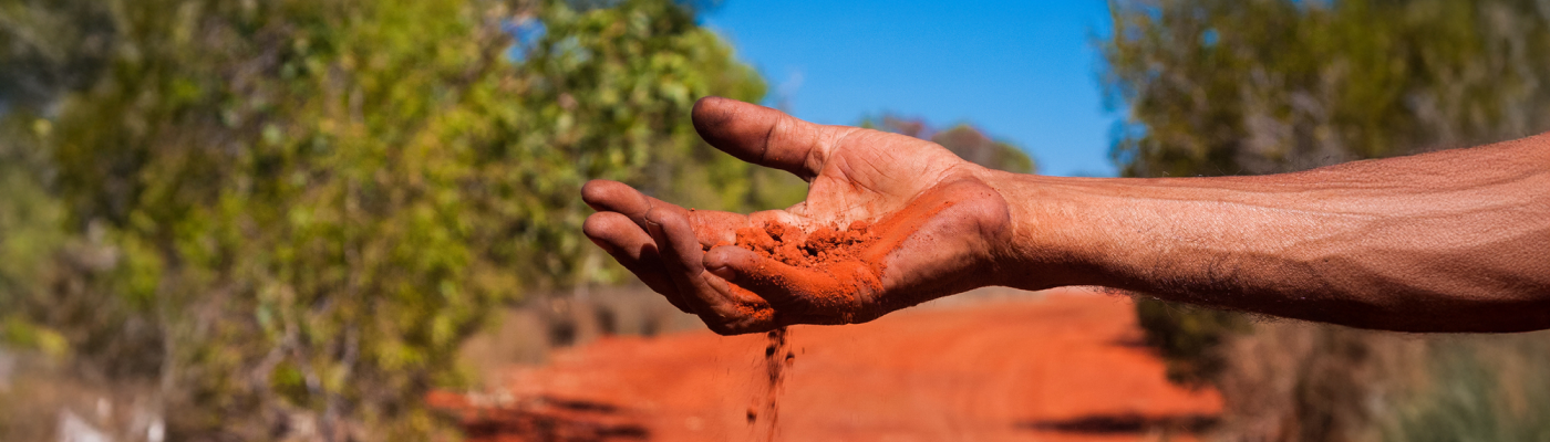 A hand sifts through vibrant red soil, letting it fall gently against a blurred backdrop of greenery and a blue sky, evoking a connection to nature.