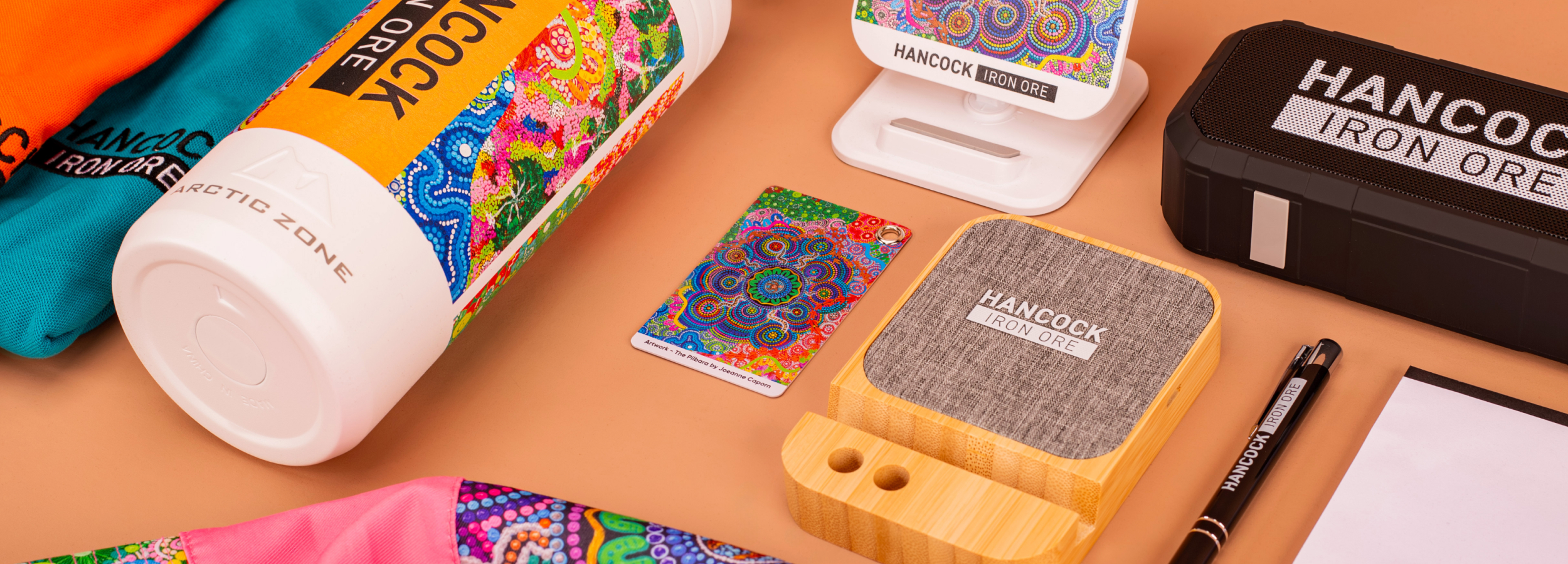 A collection of Hancock Iron Ore branded items on a tan surface, including a colourful bottle, patterned card, speaker, and accessories