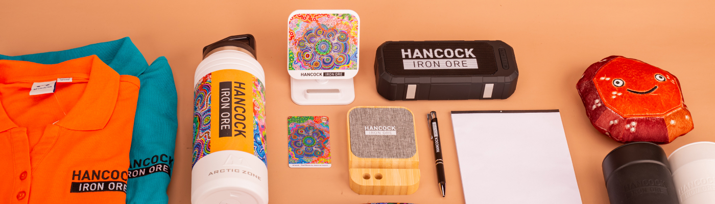 Assorted branded items on a table include a Hancock Iron Ore shirt, water bottle, speaker, colorful artwork, notepad, pen, and a smiling plush toy.