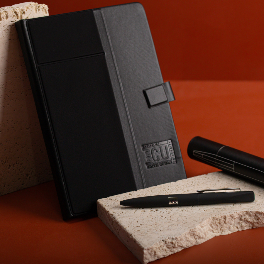 A sleek black notebook with an embossed logo leans against a textured stone.