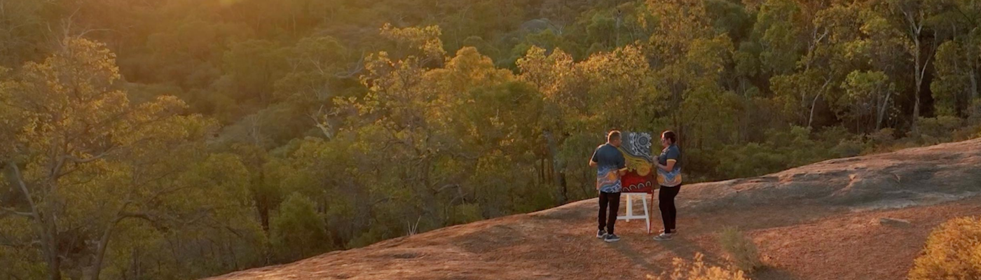 Two Aboriginal artists stand outdoors painting on a canvas mounted on an easel.