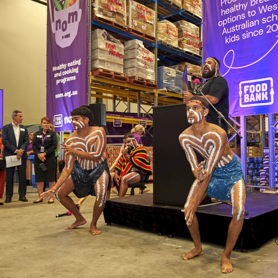 Aboriginal performers with body paint dance energetically at a Food Bank event, while a speaker addresses the audience. The mood is vibrant and cultural.