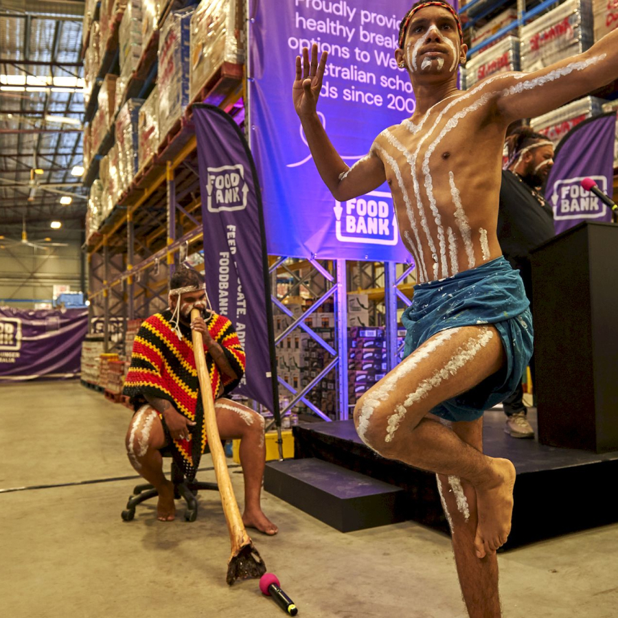 A man in traditional attire performs an Indigenous dance, marked by white body paint, while another plays a didgeridoo in a warehouse setting. Food Bank banners are in the background.