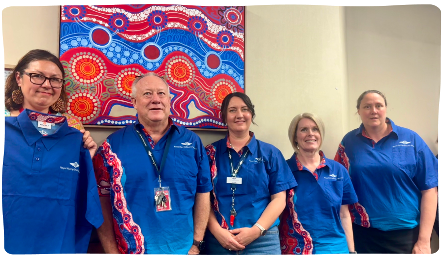 Five people in blue shirts with red and white patterns stand smiling in front of a colorful abstract painting with swirling designs and circles.