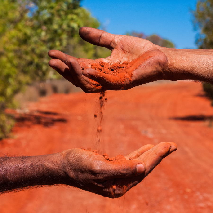 Two hands pass red soil to each other over a dirt road, surrounded by green foliage under a clear blue sky, conveying a sense of connection.