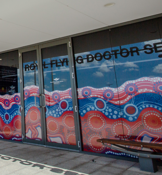 Building entrance with Royal Flying Doctor Service signage, featuring vibrant Indigenous art and acknowledgment text. Bright, welcoming atmosphere.