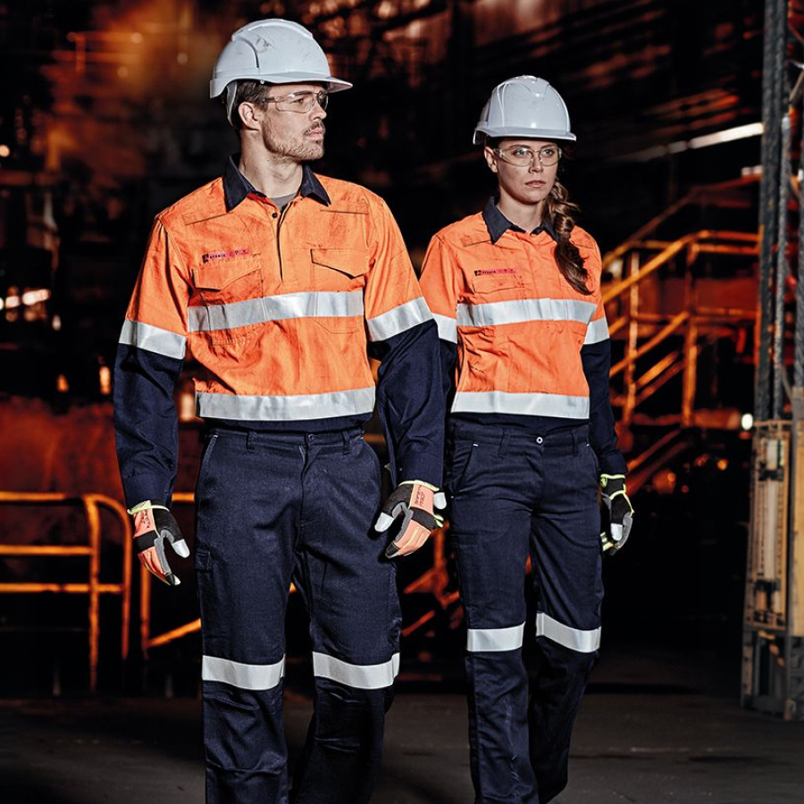 Two construction workers wearing safety gear, including bright orange reflective uniforms, helmets, and gloves, stand in an industrial setting.