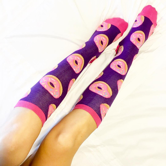 A pair of legs resting on a white bedsheet, wearing knee-high socks adorned with a pattern of pink-frosted doughnuts on a purple background