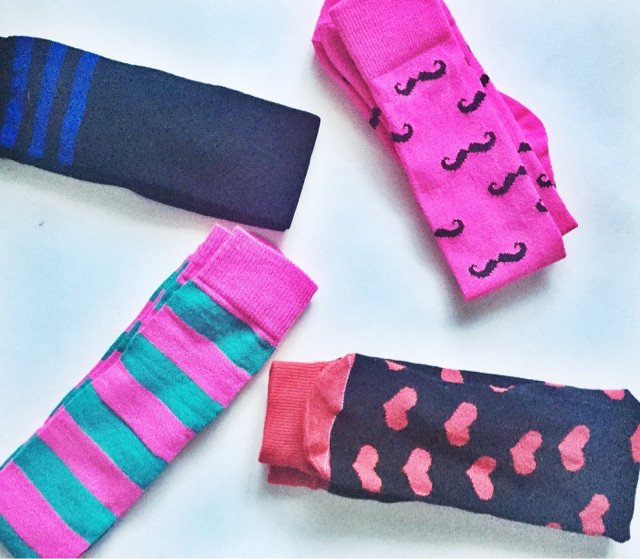 "A collection of colorful socks laid flat against a light background. From top to bottom: a black sock with blue horizontal stripes, a pink sock with black mustache patterns, a striped sock alternating between pink and teal, and a black sock with pink heart shapes.