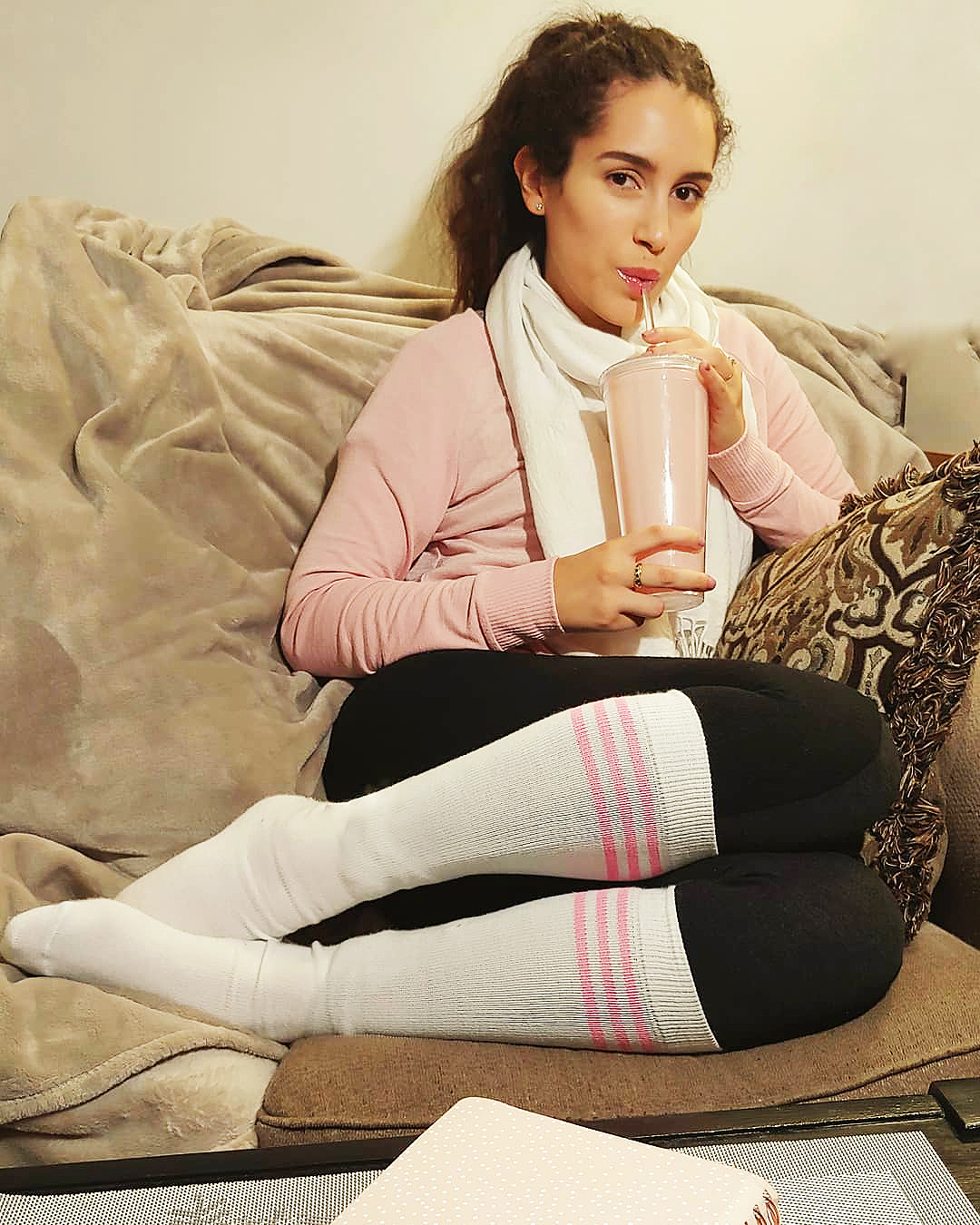 girl drinking a smoothie while sitting on the couch in leggings, long sleeve shirt, scarf and pink tube socks