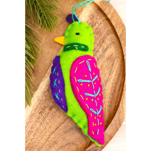 Green felt partridge ornament with pink and purple wings, turquoise and yellow embroidery, and a yellow beak, resting beside evergreen branches.